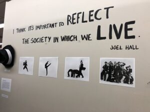 Wall text reading: "I think it's important to reflect the society in which we live. Joel Hall" with a hat and four photos displayed underneath. 