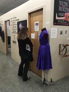 A person with brown long hair examining paper displayed on a wall while standing next to a display of a bright purple dress. 