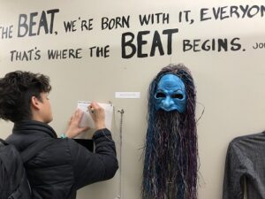 A person writing on a card against a wall next to a hung mask and wall text.
