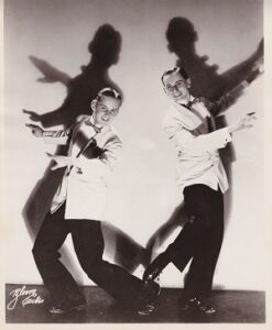 Two white men in white jackets and black pants smile toward the camera in a jazzy pose.