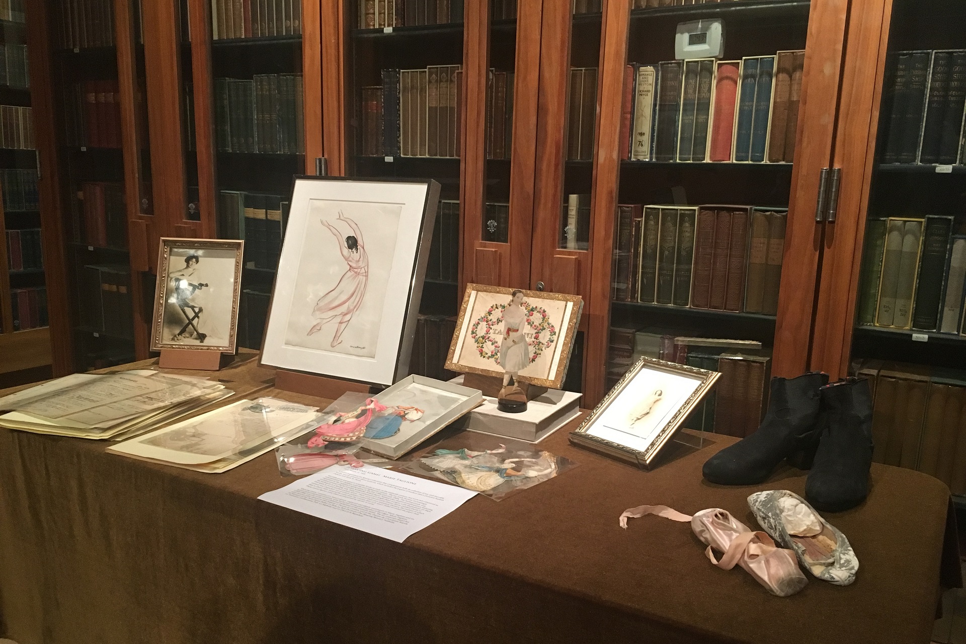 Several drawings and a pair of dance shoes on a table with a bookcase behind it