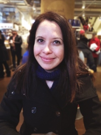 A white woman with brown hair, a black coat, and blue scarf smiles into the camera.