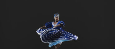 A Black woman in a royal blue dress and headdress lifts her skirt while spinning.