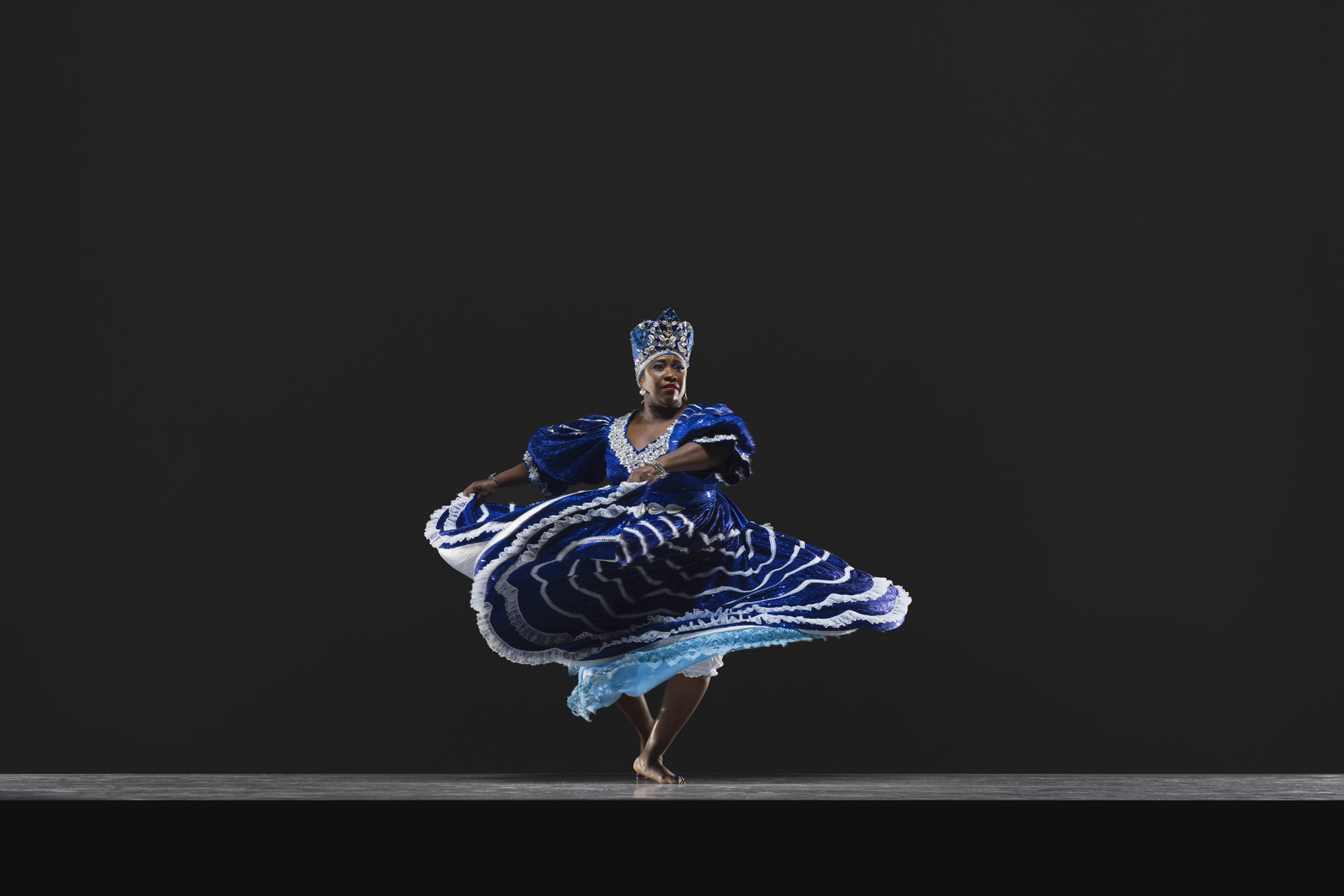 A Black woman in a royal blue dress and headdress lifts her skirt while spinning.