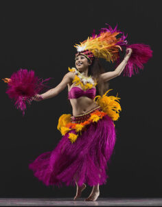 A dancer smiling in vibrant pinks and oranges performs a Polynesian dance