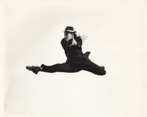 dancer in a suit and hat jumping in a split with arms crossed in front.