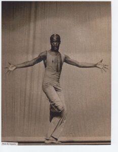 A black male dancer in a white jumpsuit with arms outstretched, fingers stretched wide.