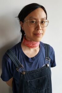 Sarah wears glasses, overalls, and a pink scarf, and looks directly at the camera.