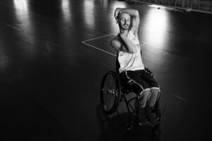 A man in a wheelchair, facing right and with his arm wrapped around his head.