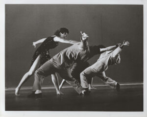 three dancers in suits reach backwards in a backbend while a fourth in a dress is in front of them reaching forward towards them.