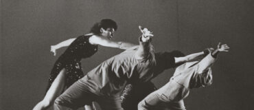 Three dancers in suits reach backwards in a backbend while a fourth in a dress is in front of them reaching forward towards them.