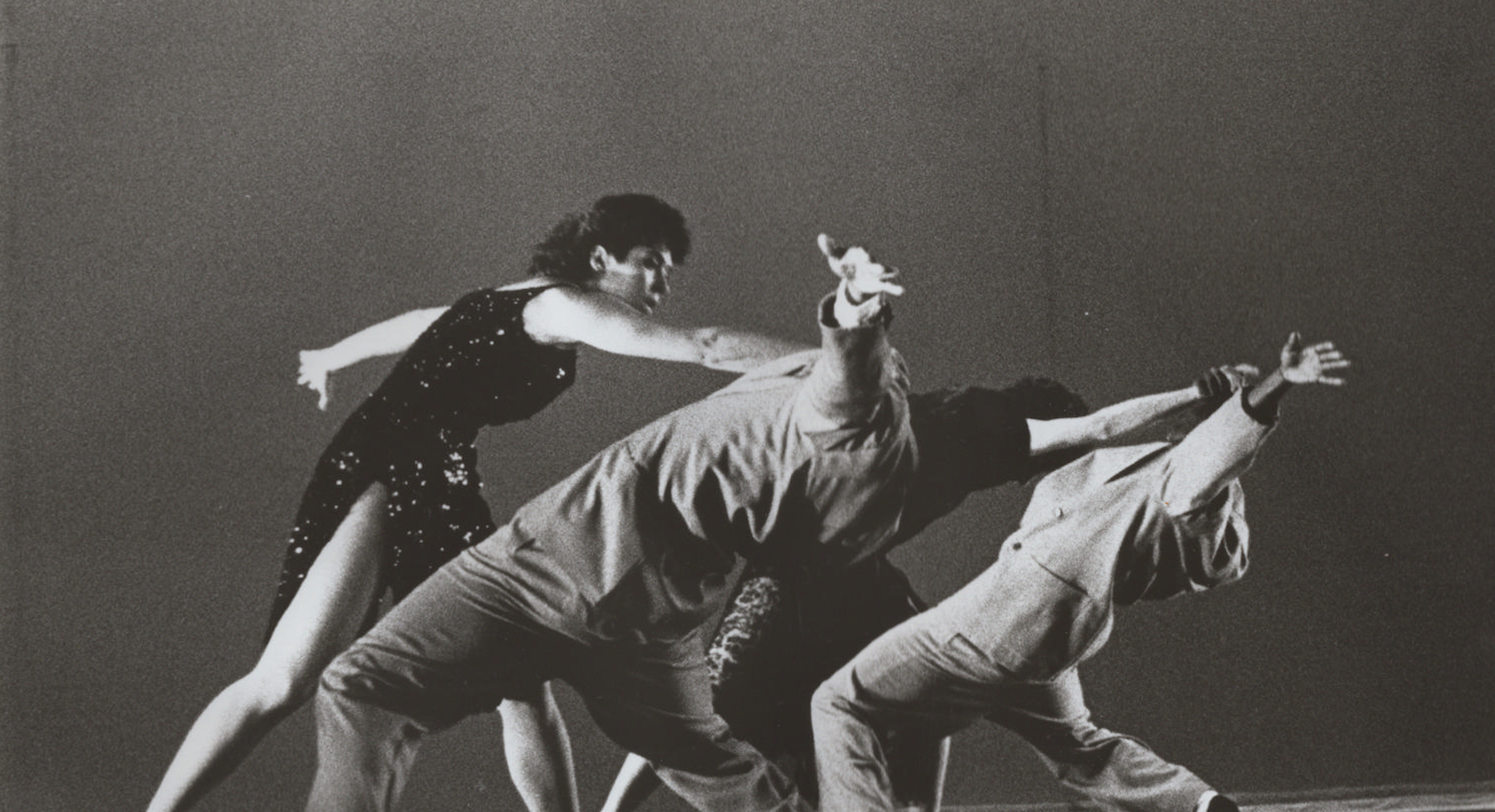 Three dancers in suits reach backwards in a backbend while a fourth in a dress is in front of them reaching forward towards them.