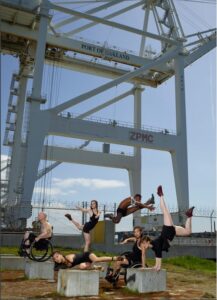 Six dancers perform in front of a large industrial crane.