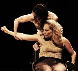 A woman sitting in a wheelchair extends her right arm, which is held by a second woman standing behind her.