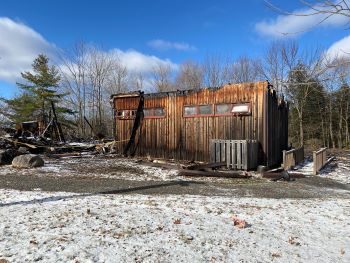 The burnt wooden walls of the theater are flanked by pine trees with snow and grass in foreground.