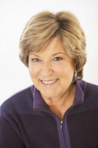 Newcomb wears a purple collared top and has short blonde hair.