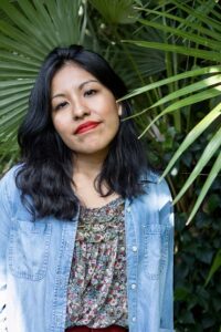 Yvette has black hair and stands in front of some tropical plants, wearing a blue shirt.