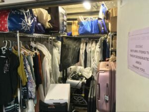 A photograph through opened doubled-doors with three steel racks of clothes hanging, bags and boxes on the top shelves, a large TV multimedia rack, a wheelchair, and a large suitcase.