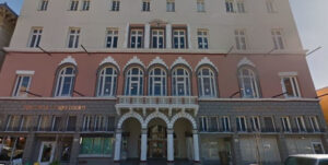 Photograph of the exterior front of a four-story building during the day time. The street-level entrance of the building opens up with three white arches surrounded by grey bricks and adjacent store window fronts to sidewalk level. The second level of the building is terracotta with narrow arched windows topped with geometric white bricks, and the third and fourth levels are painted cream with small rectangular windows.