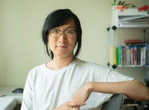 A non-binary person of Chinese descent, wearing a white top and glasses, is seated at a desk