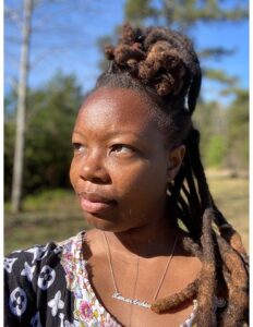 A Black woman with dreadlocks, wearing a flowered top, looks upward in an outdoor setting.