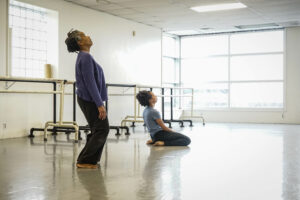 Two Black women in blue tops and dark pants in a white dance studio. One stands and one kneels while both look at the ceiling. 