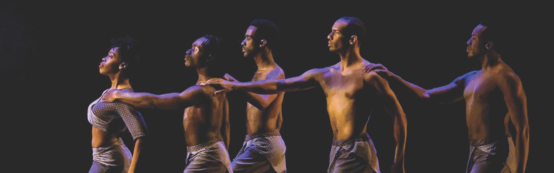 Five Black dancers stand in a line, facing left, with arms extended, against a dark background.