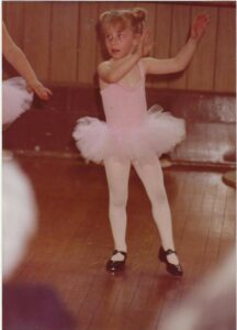 A young child in a pink leotard and tutu, pink tights, and black shoes does a dance move.