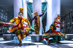 Three dancers in elaborate, colorful traditional garb dance dance among silver and white columns and platforms. 