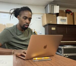 A young Black man seated at a table working on a laptop in an archive space.