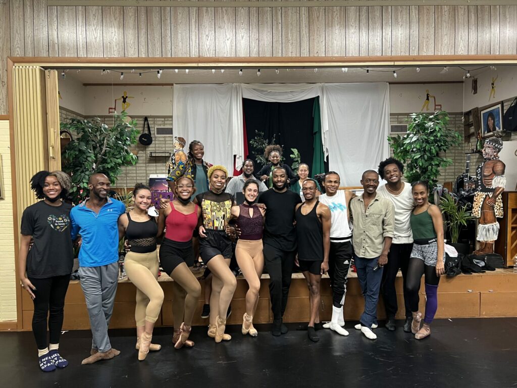 A group of people standing in a dance studio, some in dance clothes, with their arms around each other smiling.