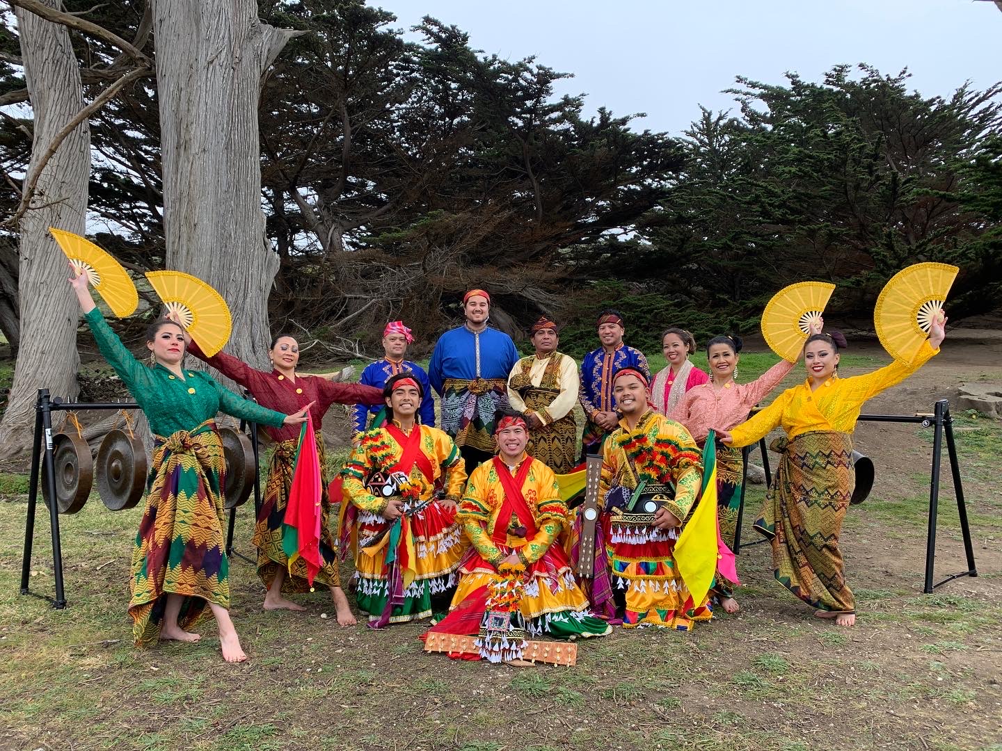 A group of 12 people in colorful garb with large fans standing outdoors on the grass in front of trees.
