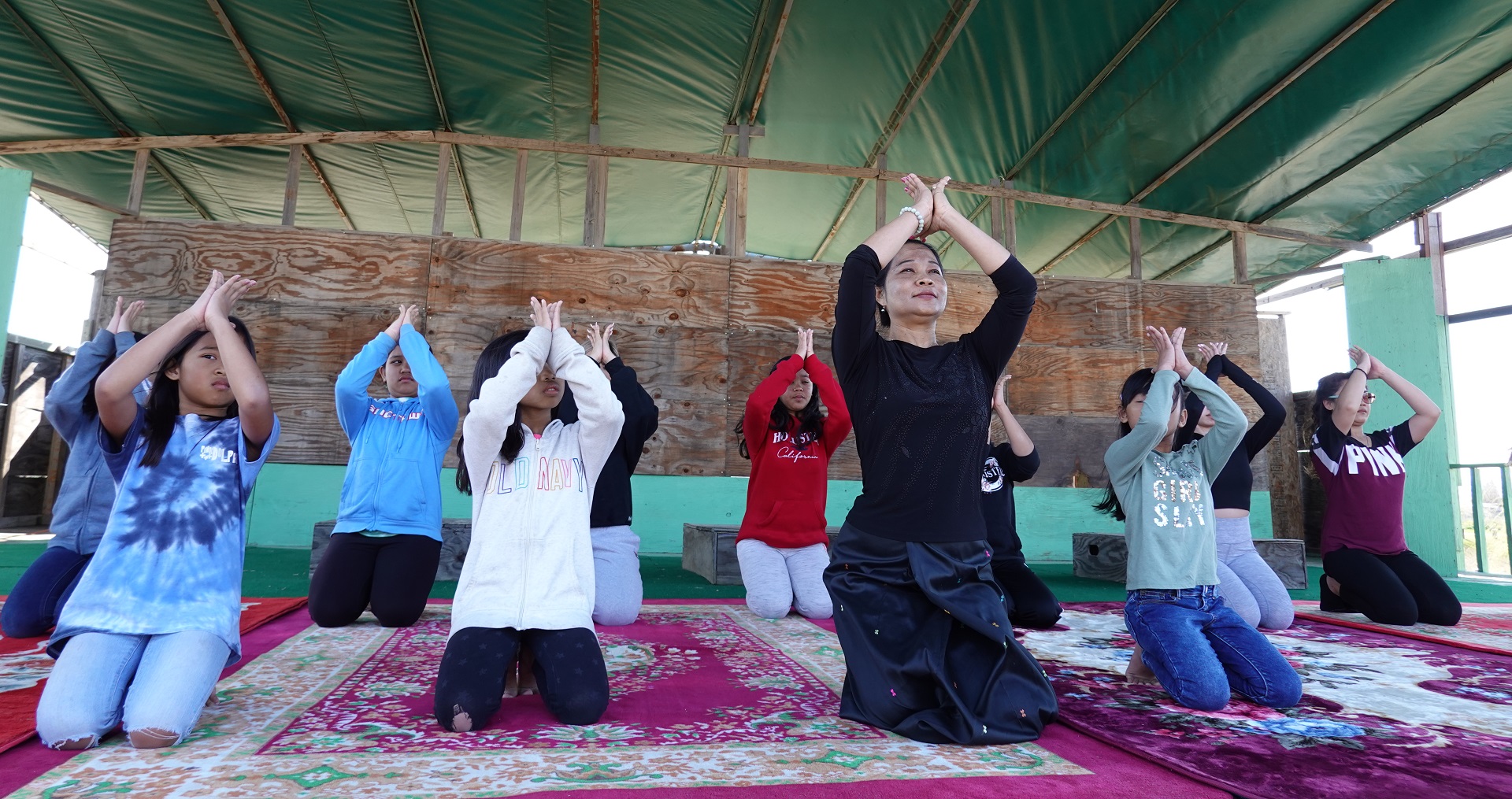 A woman and a group of children kneel on the floor performing a dance gesture