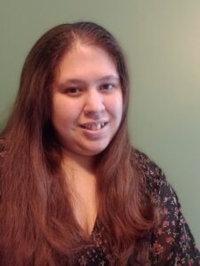 A woman with long brown hair and a flowered top looks at the camera, smiling slightly.