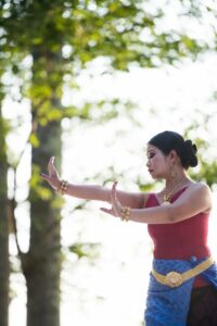 A Cambodian woman dressed in red and blue with gold jewelry dancing outdoors