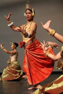 A Cambodian woman in red and gold with a gold headdress dancing, two other dancers on the floor at her sides.
