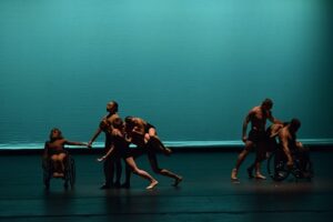 Eight dancers, two using wheelchairs, perform in front of a teal backdrop.