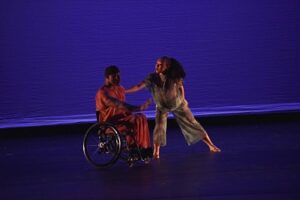 Two African-American dancers, one using a wheelchair, in front of a deep blue background.
