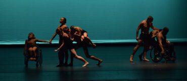Eight dancers, two using wheelchairs, perform in front of a teal backdrop.