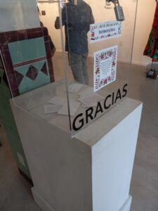 A clear box with black text reading "GRACIAS" with some slips of paper inside standing on a white pedestal. 