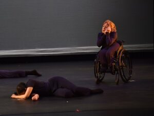Two dancers in dark costumes, one lying on the stage, the other seated in a wheelchair.