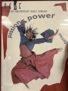 A Black female dancer dressed in a pink skirt and blue jacket leaps with arms outspread.