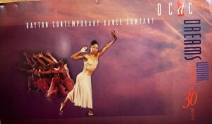 A Black female dancer in a cream-colored costume poses in front of a row of male dancers.