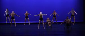Eight dancers on stage, jumping with arms outspread, against a blue backdrop.