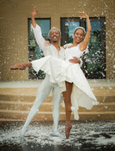 Two Black dancers dressed in white. They stand outside with snow falling around them. Their arms are around each other and they are posed balletically.