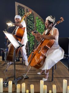 Two Black musicians dressed in white with large string instruments siting in front of a green set with candles on the floor in front of them.