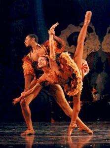 Two Black ballet dancers. The woman is on a point shoe with her leg kicked high behind her. The man behind her holds her wrists in a lunge.