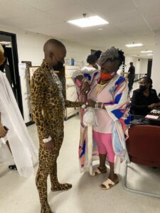 A dancer in a leapord print body suit whose costume is being adjusted by a person in pink clothes.