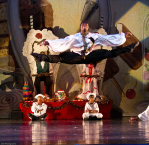A Black ballet dancer jumps high on stage with his legs and arms spread way out in a side split. People behind him sit onstage watching.