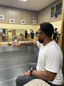 A person in a white shirt holding an arm out gesturing. Dancers in the background.